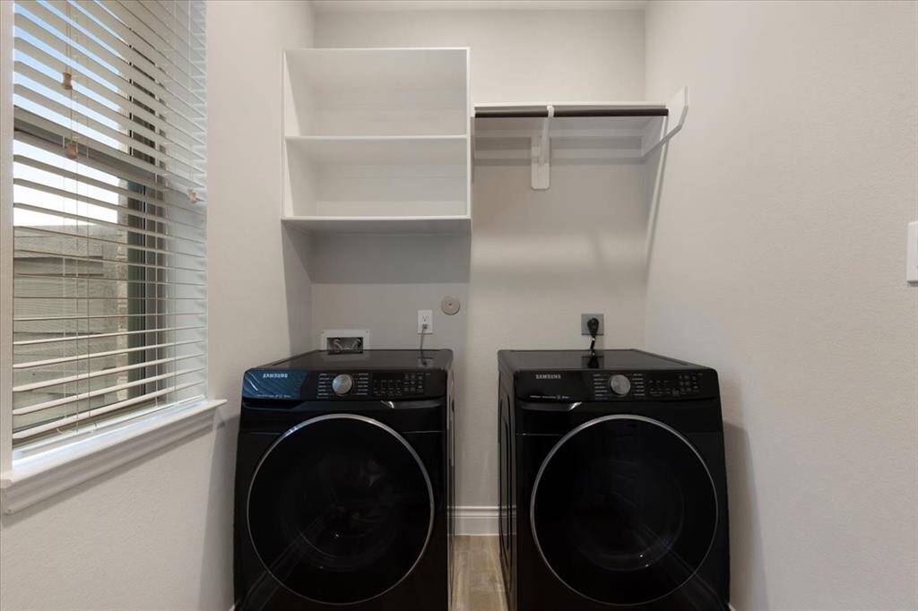 This laundry room features modern black Samsung washer and dryer units, a window for natural light, open shelving, and a hanging rod for clothes.