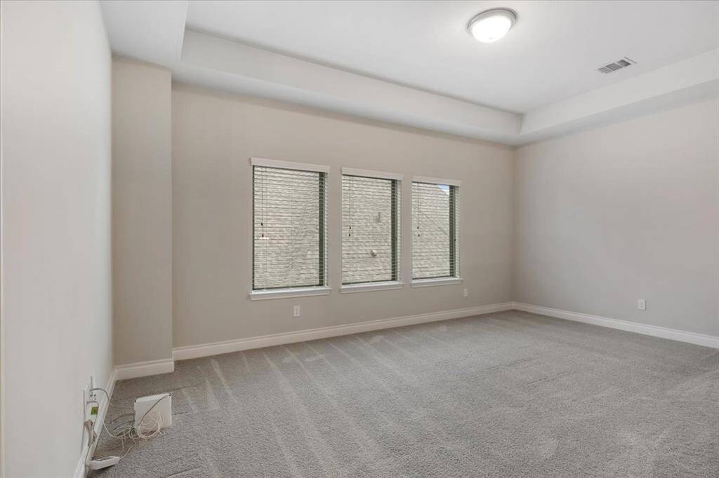 This room is spacious with neutral-colored walls and carpet, featuring three windows that allow natural light. It has a simple ceiling light fixture and minimal decor, offering a clean slate for personalization.
