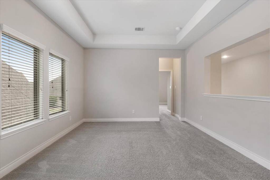 This photo showcases a bright, carpeted room with neutral walls and a tray ceiling. It features two large windows with blinds, allowing natural light to fill the space. There's an open doorway to an adjacent area, making it versatile for various uses.