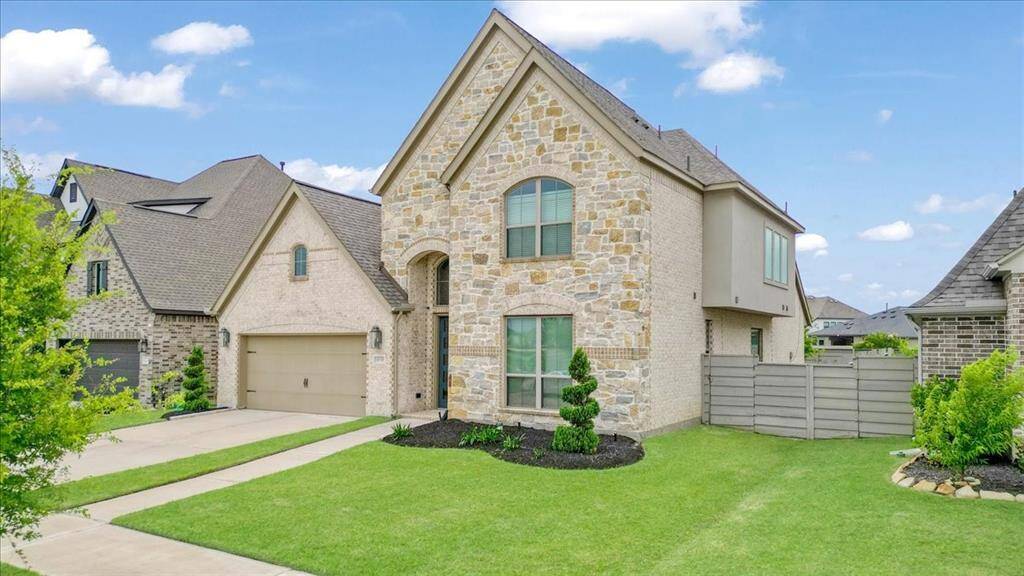 This photo showcases a modern two-story home with a stone and brick exterior, featuring a well-maintained front lawn and a two-car garage. The property is located in a suburban neighborhood with a clear, blue sky backdrop.