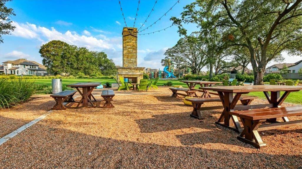 This photo showcases a welcoming outdoor community area featuring several picnic tables on a gravel surface, a central stone fireplace, and string lights overhead. The space is surrounded by lush trees, with houses visible in the background, and includes a playground, making it ideal for family gatherings and outdoor activities.