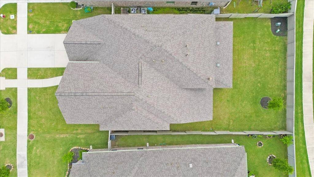 This aerial view showcases a spacious home with a well-maintained roof and a generous backyard, perfect for outdoor activities. The property is bordered by a neat driveway and lush greenery, offering privacy and a serene setting. Ideal for families seeking a peaceful neighborhood.