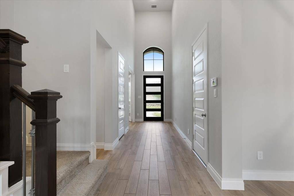This photo showcases a bright, spacious entryway with high ceilings and a modern black front door featuring glass panels. Light wood flooring complements the neutral walls, and a carpeted staircase with dark wood railing is visible to the left.