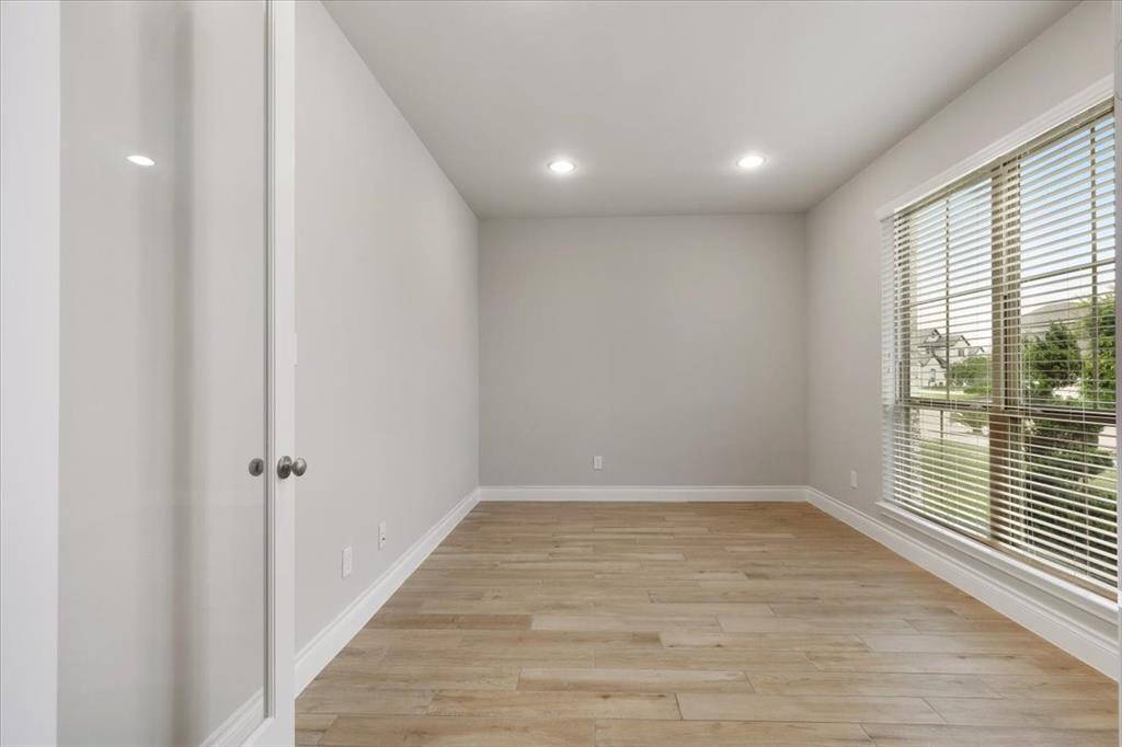 This photo showcases a bright, empty room with light wood flooring and neutral walls. It features a large window with blinds, allowing plenty of natural light, and a glass double door entrance, making it ideal for an office or study space.