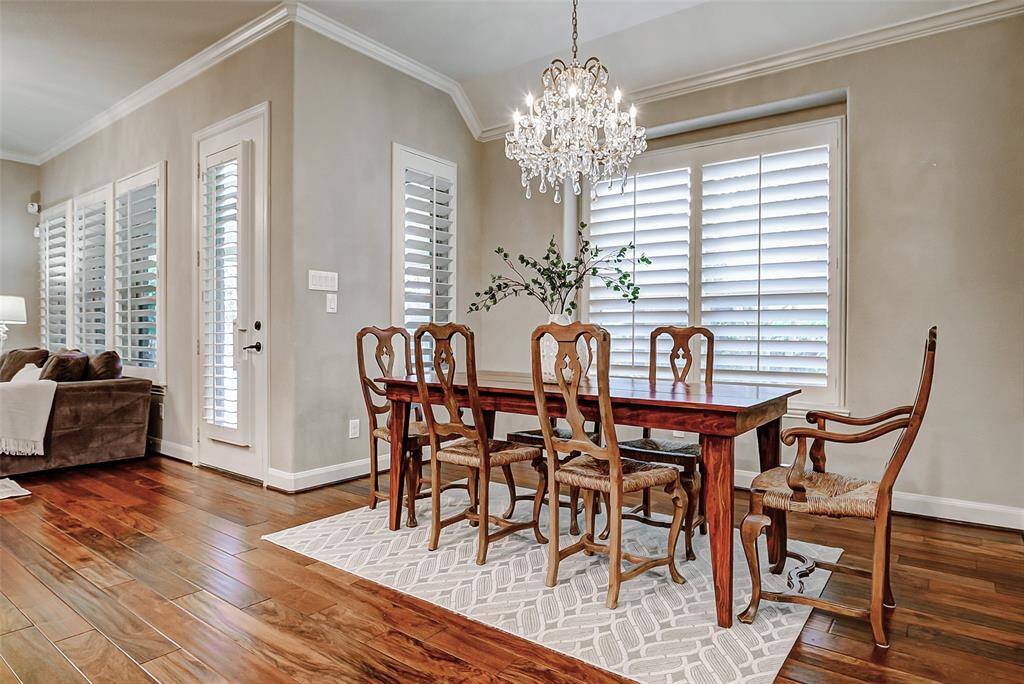 The elegant dining room features gleaming wood floors, a stunning chandelier, crown molding, and large windows with classic plantation shutters, creating sophisticated space perfect for entertaining.