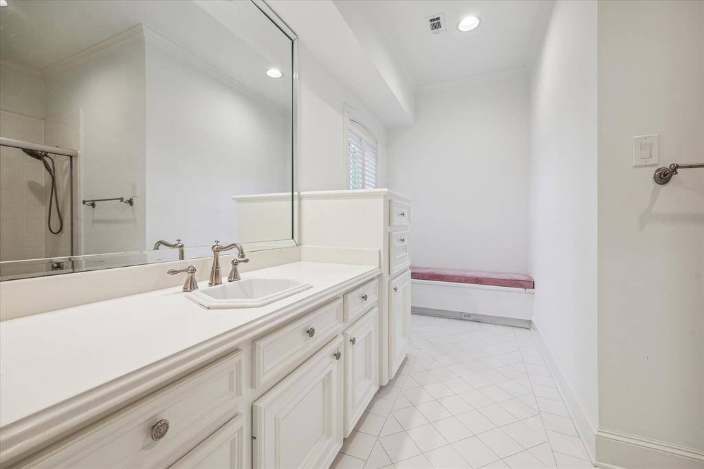 This secondary bathroom features custom beveled mirrors and sleek Corian countertops with a brushed nickel widespread faucet, complemented by a white sink and elegant white tile flooring. A tub/shower combo with a custom glass enclosure and built-in bench seat offers both style and functionality, while recessed LED lighting provides a warm, well-lit atmosphere. A window with views of the front lawn and designer neutral wall paint enhance the serene, spa- like feel of the space.