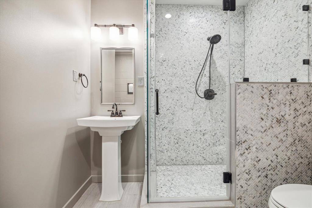 The ensuite bathroom features a pedestal sink with a brushed nickel faucet, complemented by a designer polished nickel mirror and brushed nickel sconce lighting. The walk-in shower with marble floor and wall tiles, designer neutral tile flooring, and recessed LED lighting create a luxurious yet functional space, with convenient access to the media room and bedroom.