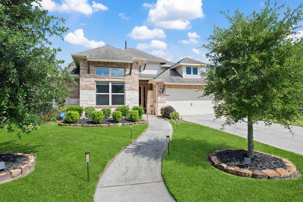 This is a charming single-story brick home with a manicured front yard, featuring lush green grass and neatly trimmed bushes. A curved walkway leads to the inviting entrance, and there's a two-car garage on the side. The house has a modern, welcoming appearance in a suburban setting.