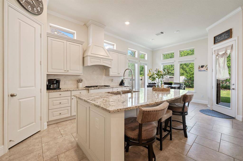 This bright, airy kitchen features elegant white cabinetry, a large granite island with seating, and stainless steel appliances. The space opens to a lush backyard through glass doors and windows, enhancing the inviting atmosphere.