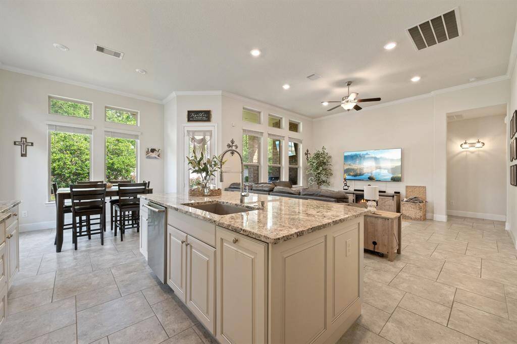 This open-concept living area features a spacious kitchen with a large island and granite countertops. The adjacent breakfast area is bright with natural light from large windows. The living room is cozy with a ceiling fan and ample space, perfect for relaxation or entertaining. The neutral tile flooring and modern finishes create a welcoming atmosphere.