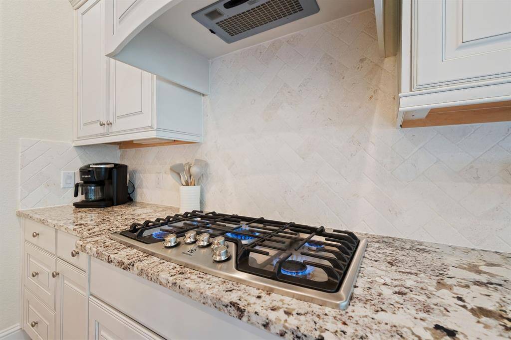 This kitchen features elegant white cabinetry and a sleek granite countertop with a modern gas stove. The backsplash is a stylish herringbone tile design, and there's a built-in range hood.