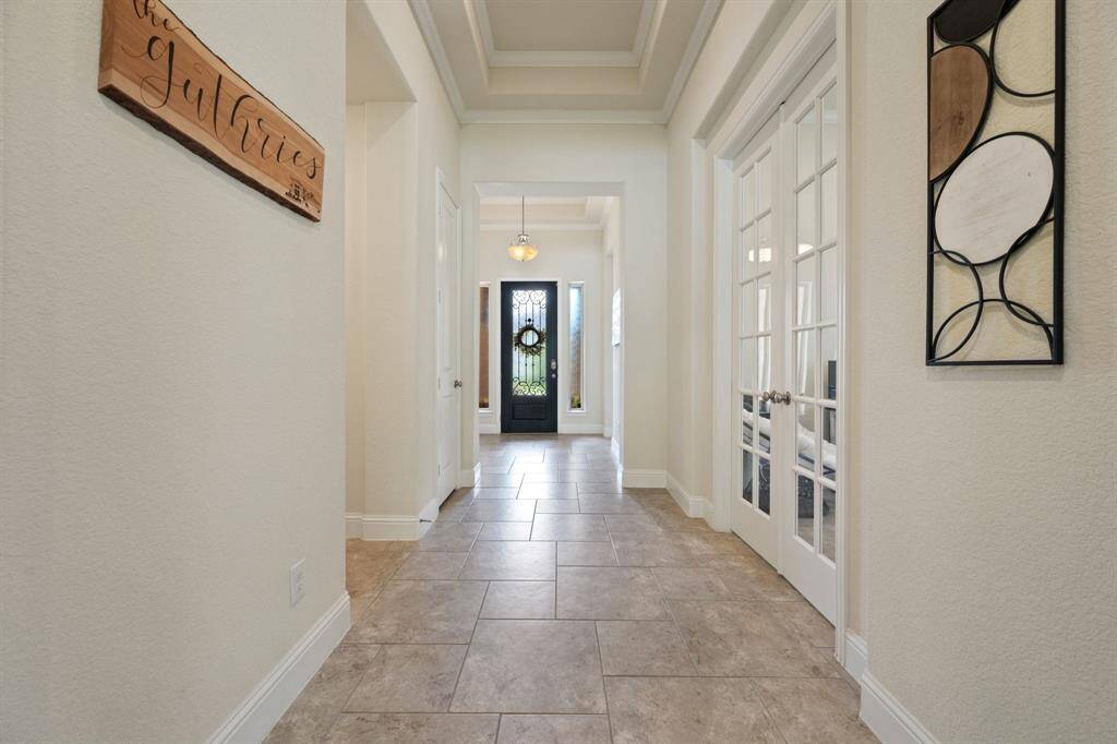 This photo features a bright, welcoming foyer with tiled flooring, leading to a front door adorned with glass and ironwork. The space includes French doors opening to a office room, offering a spacious and elegant entryway ideal for a modern home.
