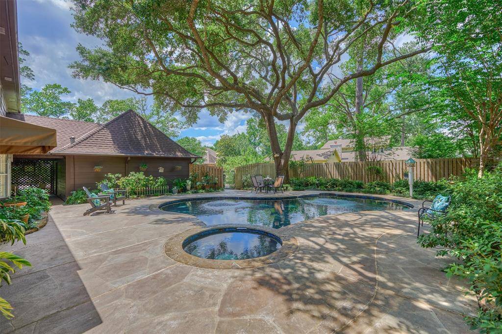 This serene backyard features a large, inviting pool surrounded by lush greenery and mature trees, offering ample shade and privacy. The patio area provides space for relaxation and outdoor seating, perfect for entertaining.