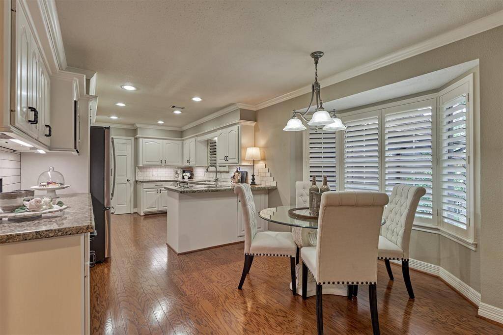 This photo showcases a bright, elegant kitchen with white cabinetry, granite countertops, and hardwood flooring. It features a cozy dining area, complemented by large windows with plantation shutters that provide ample natural light.