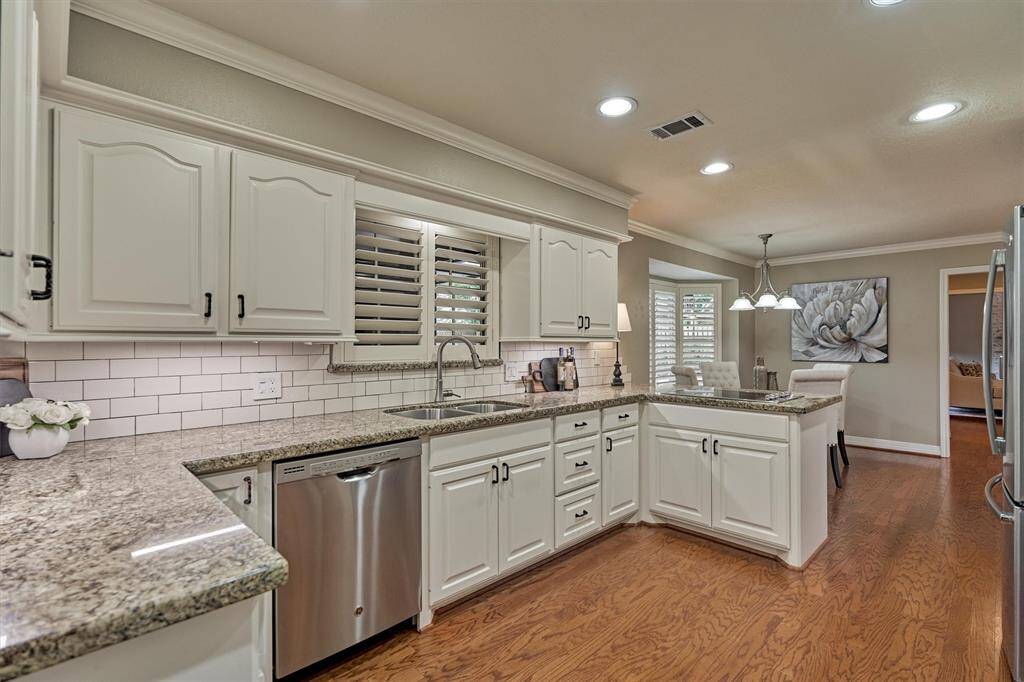 The large space is well-lit with recessed lighting and has an open layout leading to a dining area, enhanced by warm wood flooring and a stylish backsplash.