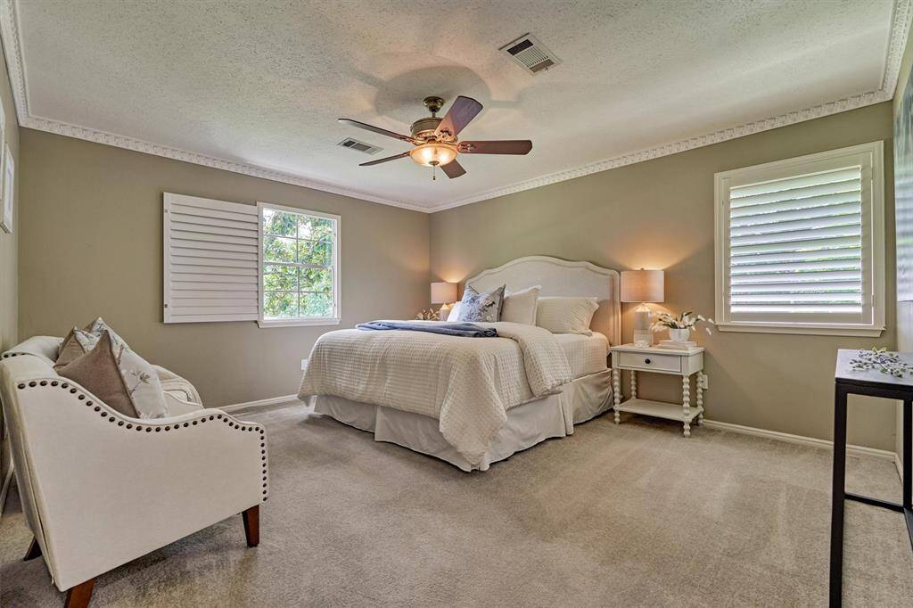 Bright and inviting primary bedroom with neutral tones, elegant ceiling fan, and ample natural light from two shuttered windows. Perfect for relaxation and comfort.