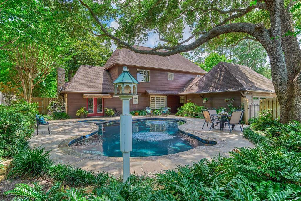 This photo showcases a charming backyard with a pool surrounded by lush greenery and a large, shady tree. The house features a warm, rustic exterior with multiple rooflines, and there's a cozy patio area with outdoor seating, perfect for relaxing or entertaining.