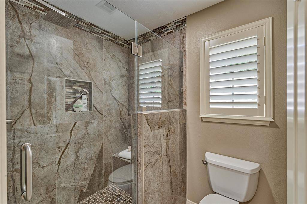 This bathroom features a sumptuous & modern shower with elegant stone tile, a built-in niche for toiletries, and a glass enclosure. It also includes a window with shutters, offering a clean and stylish design.