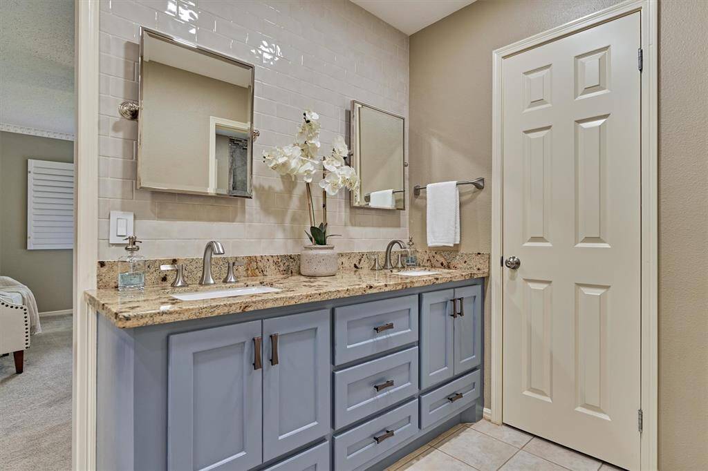 Stylish double vanity with granite countertops and blue cabinetry. It includes dual sinks, modern fixtures, and a decorative backsplash. The space is bright and inviting, with neutral-toned walls and tile flooring, offering a clean and contemporary look.