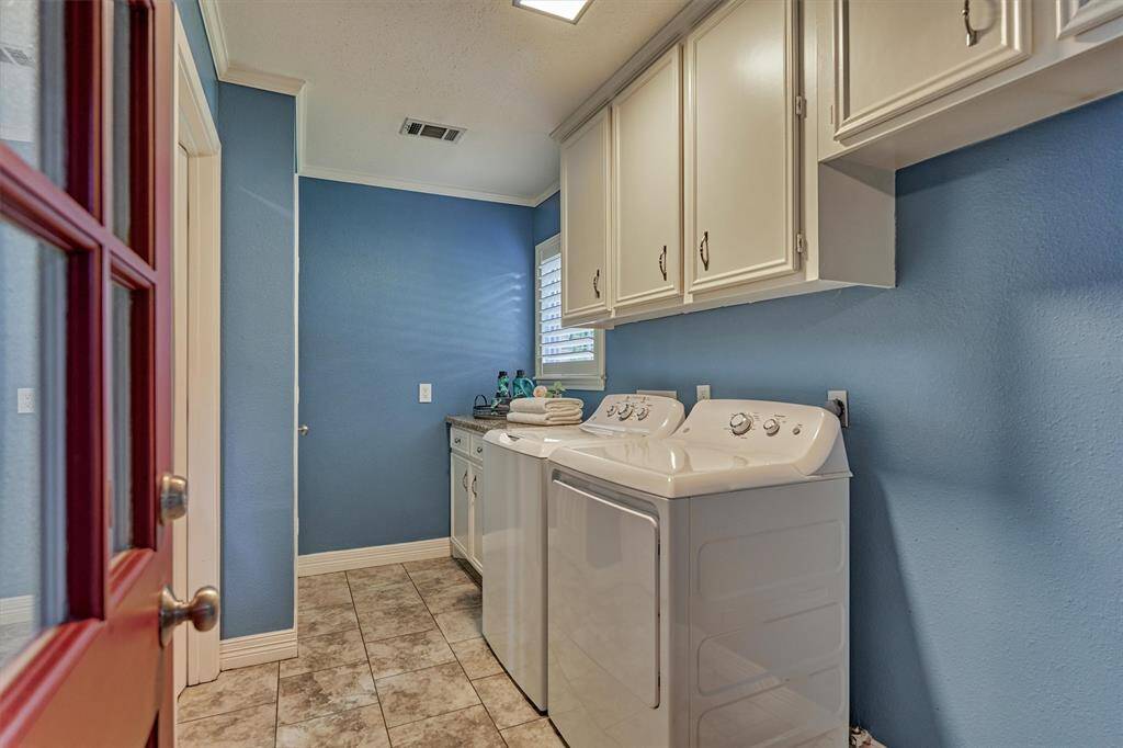 This photo showcases a well-lit laundry room with soothing blue walls, ample overhead cabinetry for storage, tiled flooring, a cleaning closet (not seen) and a red door, offering both functionality and style.