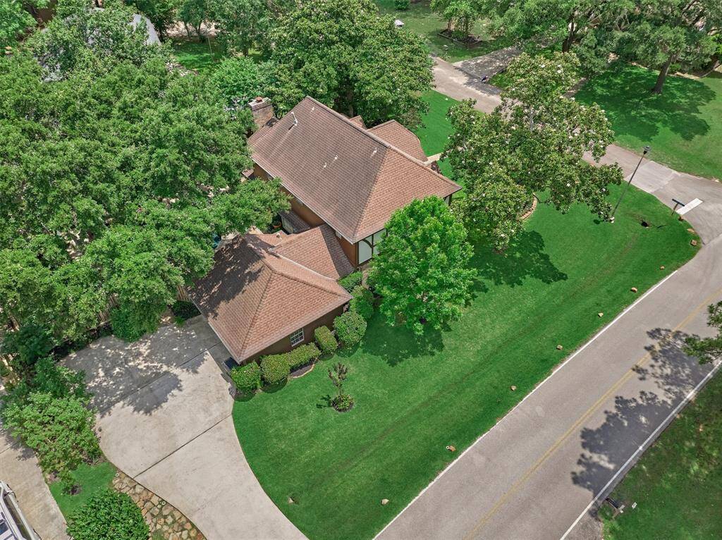 This aerial photo showcases a charming home with a sloped roof, surrounded by lush greenery and mature trees. It features a well-maintained lawn, a spacious driveway, and is located on a quiet, tree-lined street.
