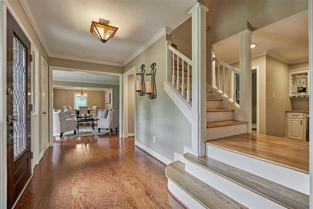 This inviting home features a bright entryway with warm wood flooring leading to a cozy living area. A staircase with elegant white railings adds charm, and there's a glimpse of a modern entertaining bar. Stylish lighting and neutral tones throughout create a welcoming atmosphere.