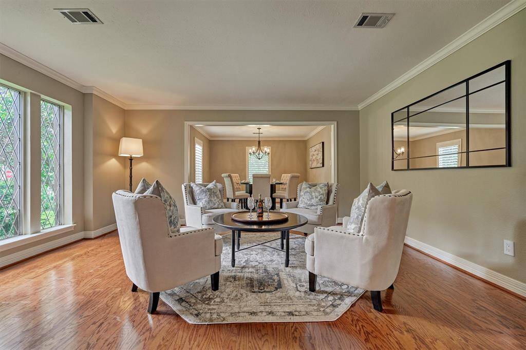 This inviting living room features warm wood flooring, elegant neutral walls, and large windows for natural light.  A mirror and tasteful lighting enhance the room's charm, while an open doorway leads to a dining area, offering a seamless flow for entertaining.