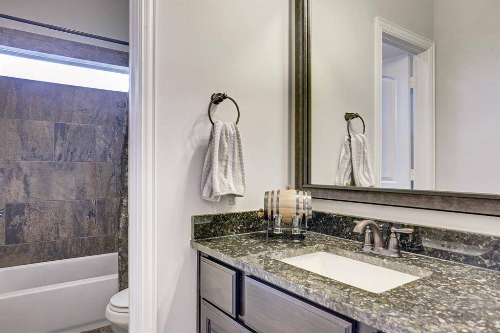This bathroom features a sleek design with a granite countertop, modern fixtures, and a large mirror. The shower area has stylish stone tiles and a built-in bathtub, creating a contemporary and functional space.