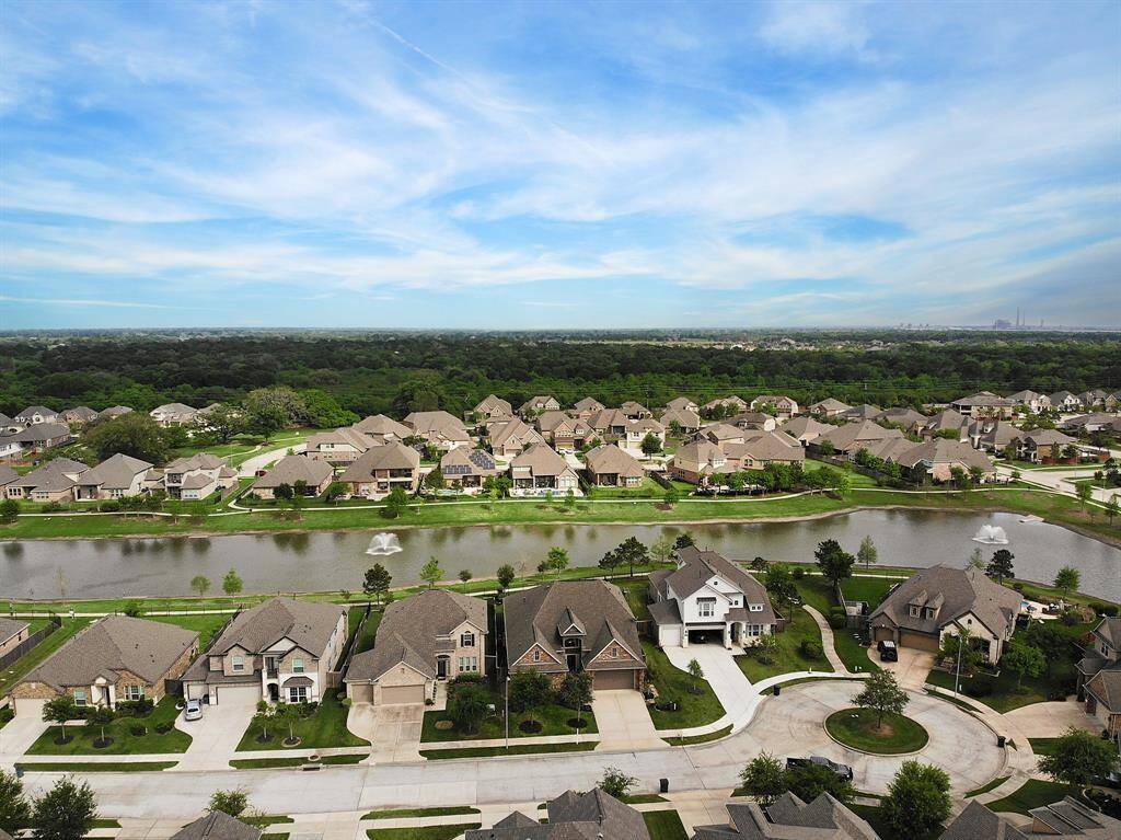 This photo showcases a picturesque suburban neighborhood with well-maintained homes, tree-lined streets, and a serene pond featuring fountains. The area offers a tranquil setting with lush greenery and a spacious, family-friendly environment.