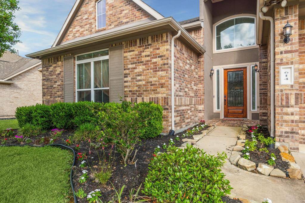 Charming brick home with a welcoming front entry, featuring a wooden door and decorative glass. The well-maintained landscaping includes lush bushes and colorful flowers, enhancing curb appeal. A paved walkway leads to the inviting porch, creating a warm and attractive entrance.