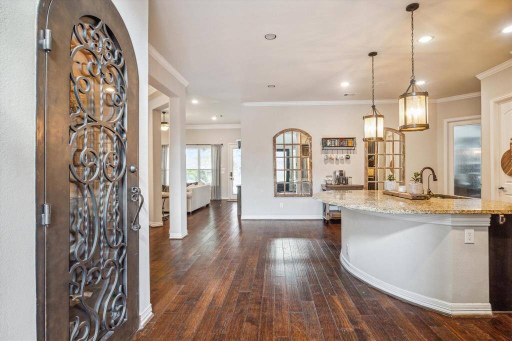 Greeted by a beautifully designed entryway and kitchen. This photo showcases a welcoming entryway with a decorative iron door and hardwood floors leading into an open-concept living area. The kitchen features a curved granite countertop with pendant lighting, and there's ample natural light from large windows, creating a warm and inviting space.