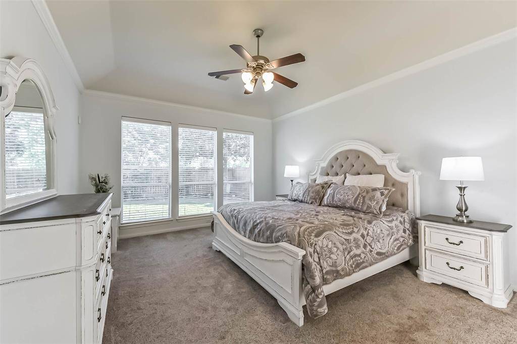 Primary suite offers a relaxing getaway. It has a cozy carpeted floor, soft neutral tones, and ample natural light from three large windows. A ceiling fan adds comfort to the airy, inviting space.