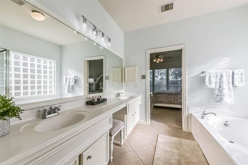 Bright and spacious bathroom with double sinks, a large mirror, and ample natural light. Features a luxurious soaking tub and connects to a bedroom, enhancing convenience and style.