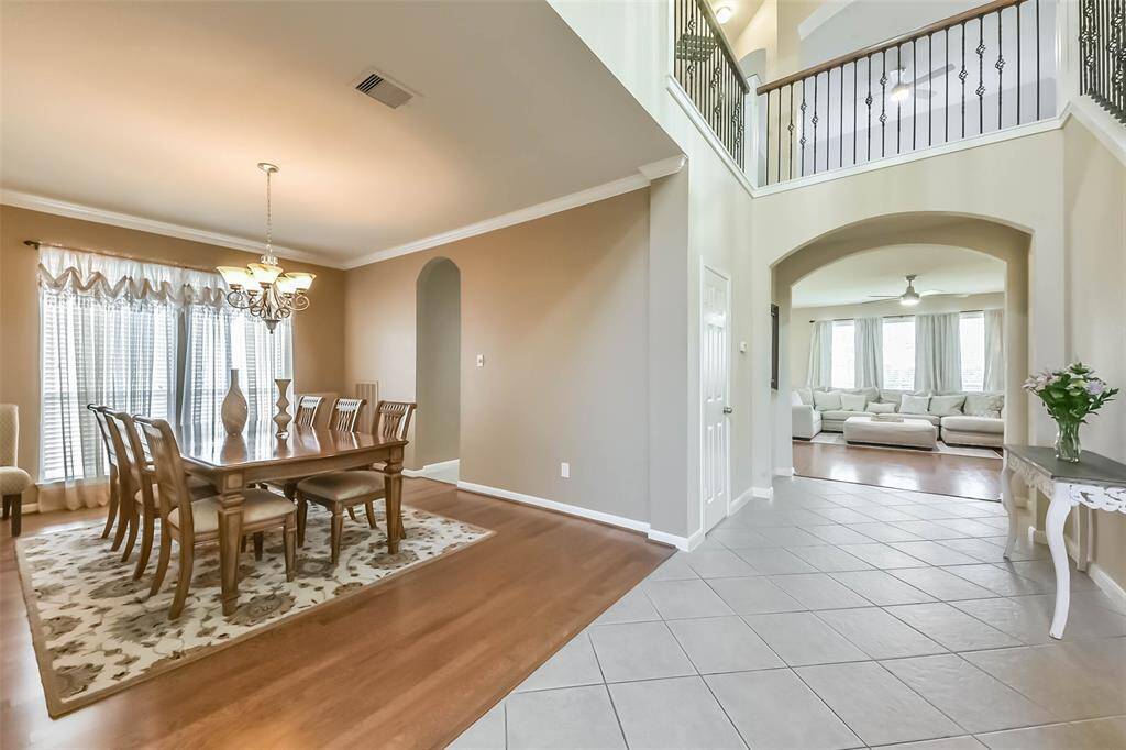 This photo showcases a spacious, open-concept living area with a formal dining room featuring elegant wood flooring and a chandelier. The adjacent living room has a cozy, inviting feel with ample natural light from large windows. The entryway is bright with tiled flooring and a high ceiling, enhancing the home's airy ambiance.
