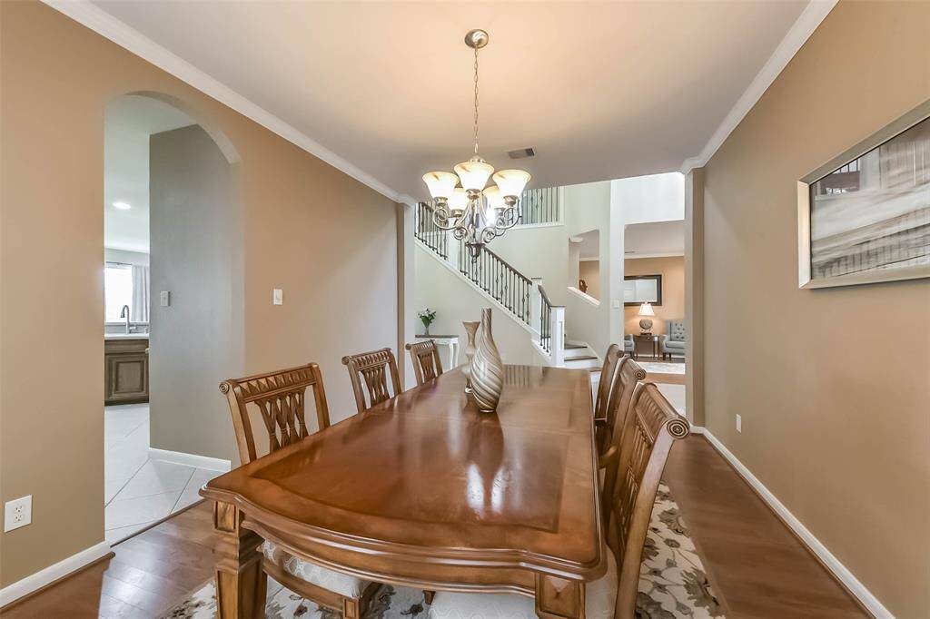 This photo showcases a spacious dining area with a wooden table and elegant chairs, lit by a stylish chandelier. The room opens up to a staircase and a bright, airy living space, creating a welcoming and connected environment.