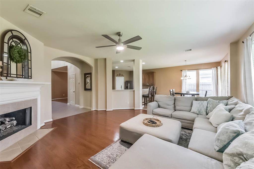 This spacious living area features wood flooring, a cozy fireplace, and room for a large sectional sofa. It opens to a dining area with ample natural light and a view into the kitchen with a breakfast bar. The neutral tones create a warm and inviting atmosphere.