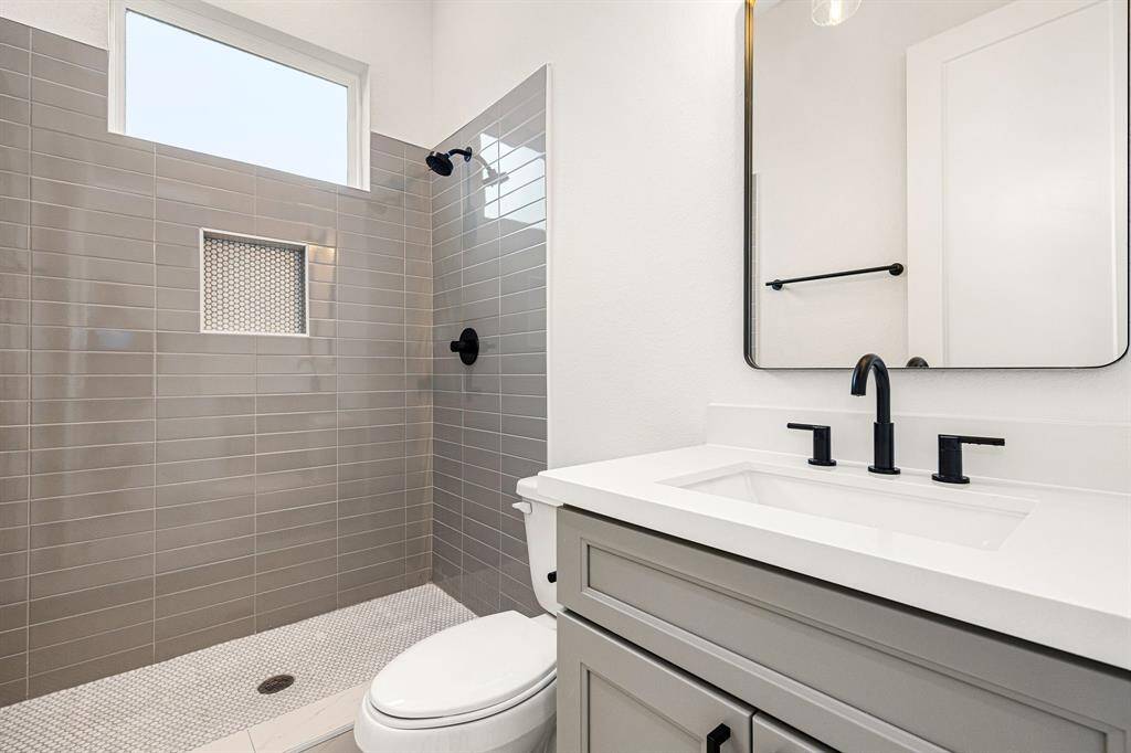 The secondary bathroom boasts elegant light countertops paired with contemporary grey cabinetry, creating a refined and airy atmosphere. The oversized walk-in shower, with its custom tile surround, offers a spa-like experience, while the decorative mirror adds a touch of sophistication. The sleek fixtures and modern finishes complement the tile floors, enhancing the bathroom’s sleek design.