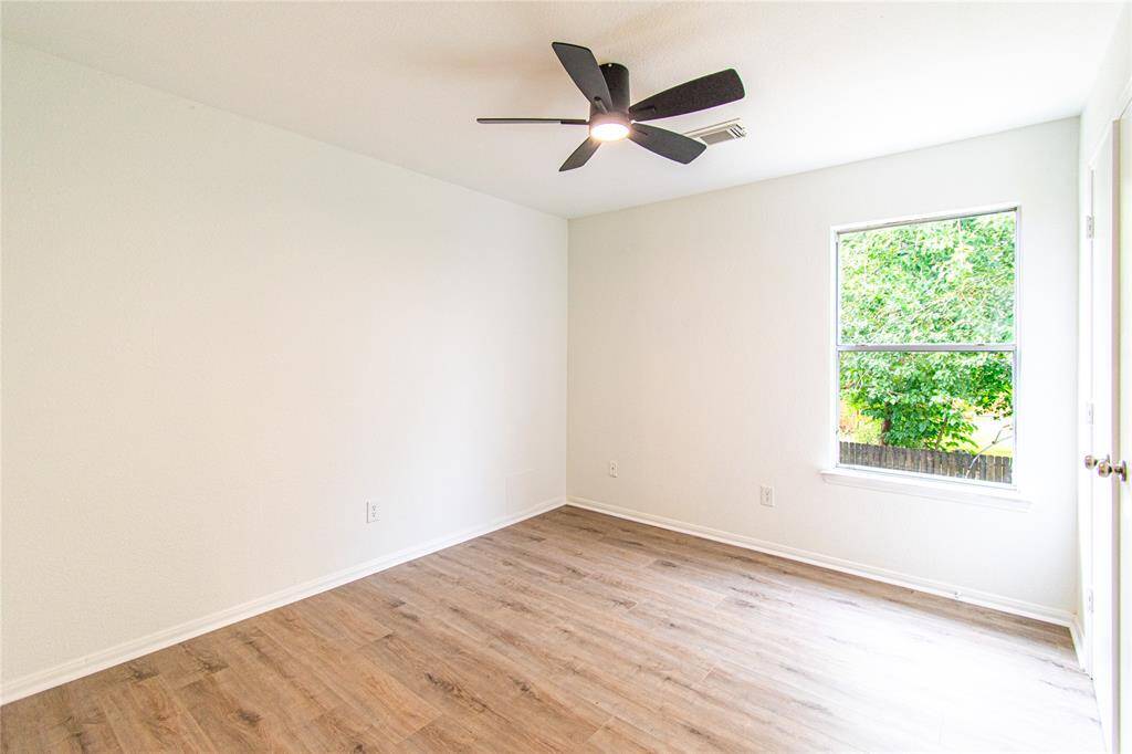 This bright, airy room features fresh white walls, a stylish ceiling fan, and a window offering a view of greenery. The space is enhanced by wood-look flooring, creating a warm and inviting atmosphere.