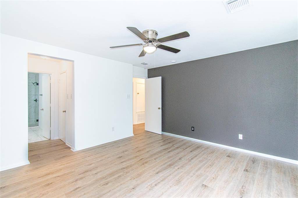 This spacious bedroom features a modern ceiling fan, light wood flooring, and a chic dark accent wall. It connects to a bathroom with a tiled shower, offering a clean and contemporary look.
