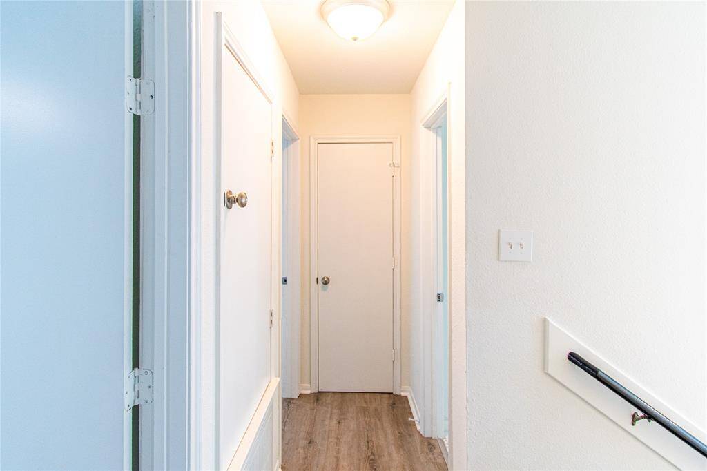 Hallway with light-colored walls and wood flooring. It features multiple doors and a staircase with a simple handrail. The space is well-lit with a ceiling light fixture.