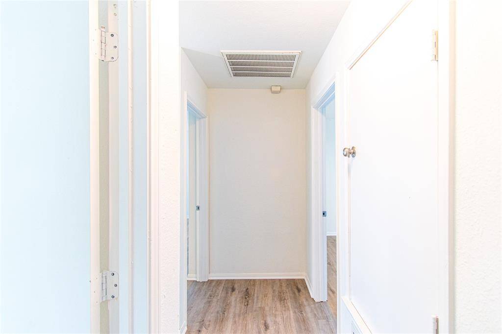 Hallway with light-colored walls and wood flooring. There are several doors leading to different rooms, and an air vent is visible on the ceiling.