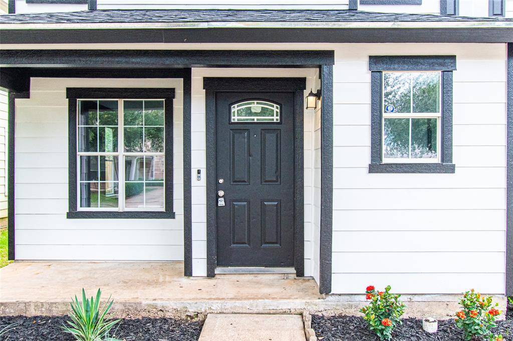 Charming entryway with a black front door and matching window frames, set against white siding. Features a small porch and a neat garden area with plants and flowers.