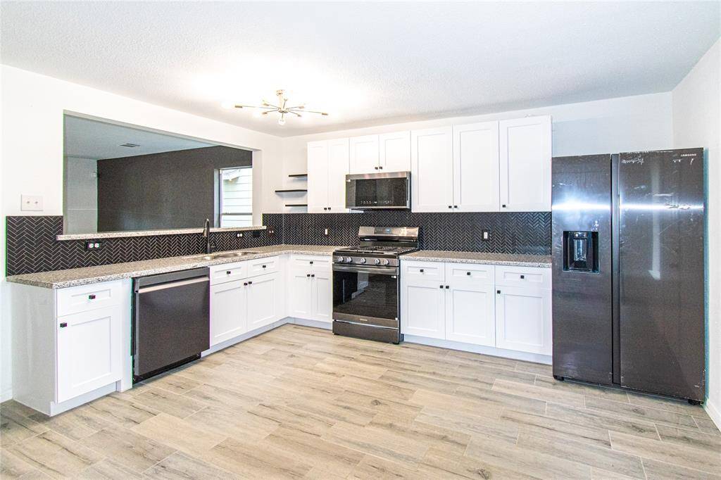 Modern kitchen features sleek black appliances, white cabinetry, and a stylish herringbone tile backsplash. It has ample countertop space, a gas stove, and a large refrigerator. The open layout and light wood flooring enhance the bright, spacious feel.