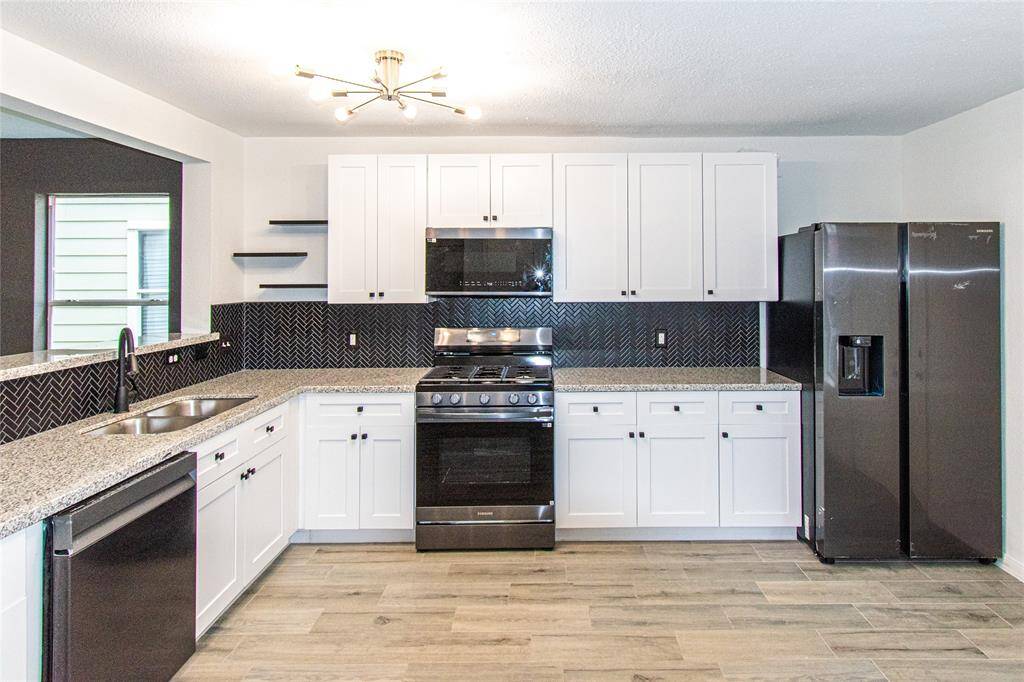 Modern kitchen with sleek white cabinetry and granite countertops. Features a stainless steel gas stove, microwave, and a double-door refrigerator. The stylish black herringbone backsplash adds a touch of elegance, complemented by wood-look tile flooring and contemporary lighting.