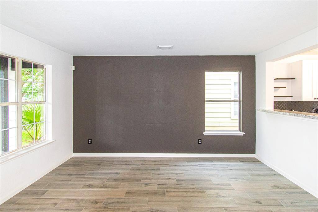 Bright living area with wood-like tile flooring, neutral walls, and a dark accent wall. Features large windows for natural light and an open view into the kitchen.