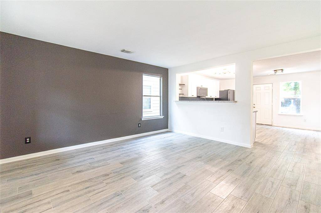 Open living space with light wood floors and a modern feel. The room features a dark accent wall, a window, and an open kitchen area with a bar counter, leading into another room with additional windows.