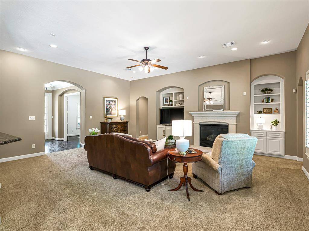 The gas log fireplace, framed by an elegant cast-stone surround, serves as a striking focal point, and the archways at the shelving and art niche echo the room’s graceful design elements, adding to its charm.