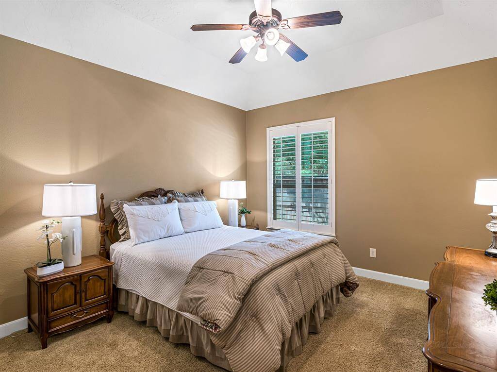 This spacious secondary bedroom features a large walk-in closet, plantation shutters, plush carpet, 5-inch baseboards, and a high ceiling with a fan—offering comfort and style.