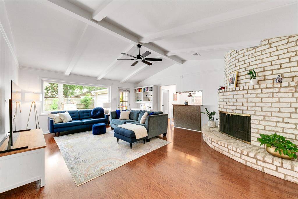 This spacious living room features a vaulted ceiling with exposed beams, a cozy white brick fireplace, and built-in shelving.