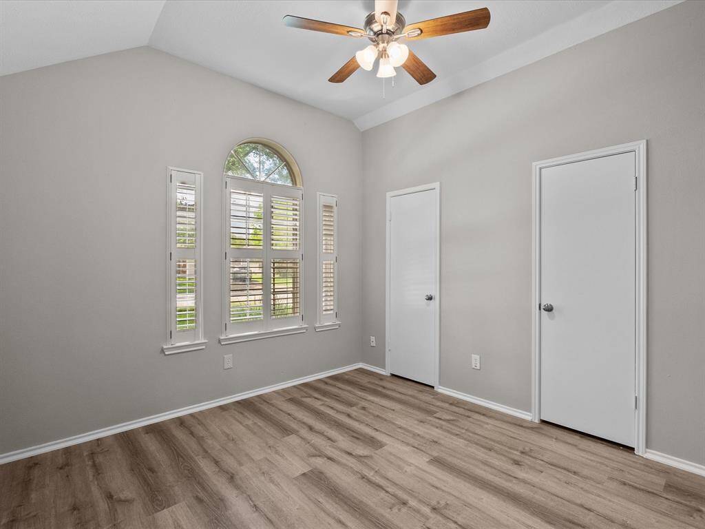Second bedroom features a vaulted ceiling with a ceiling fan, neutral walls, and wood-look flooring. It has a large arched window with plantation shutters, and two closet doors, offering a bright and airy feel.